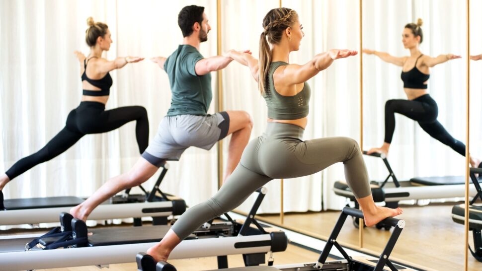 Two women in a Pilates reformer class doing standing leg and core exercises